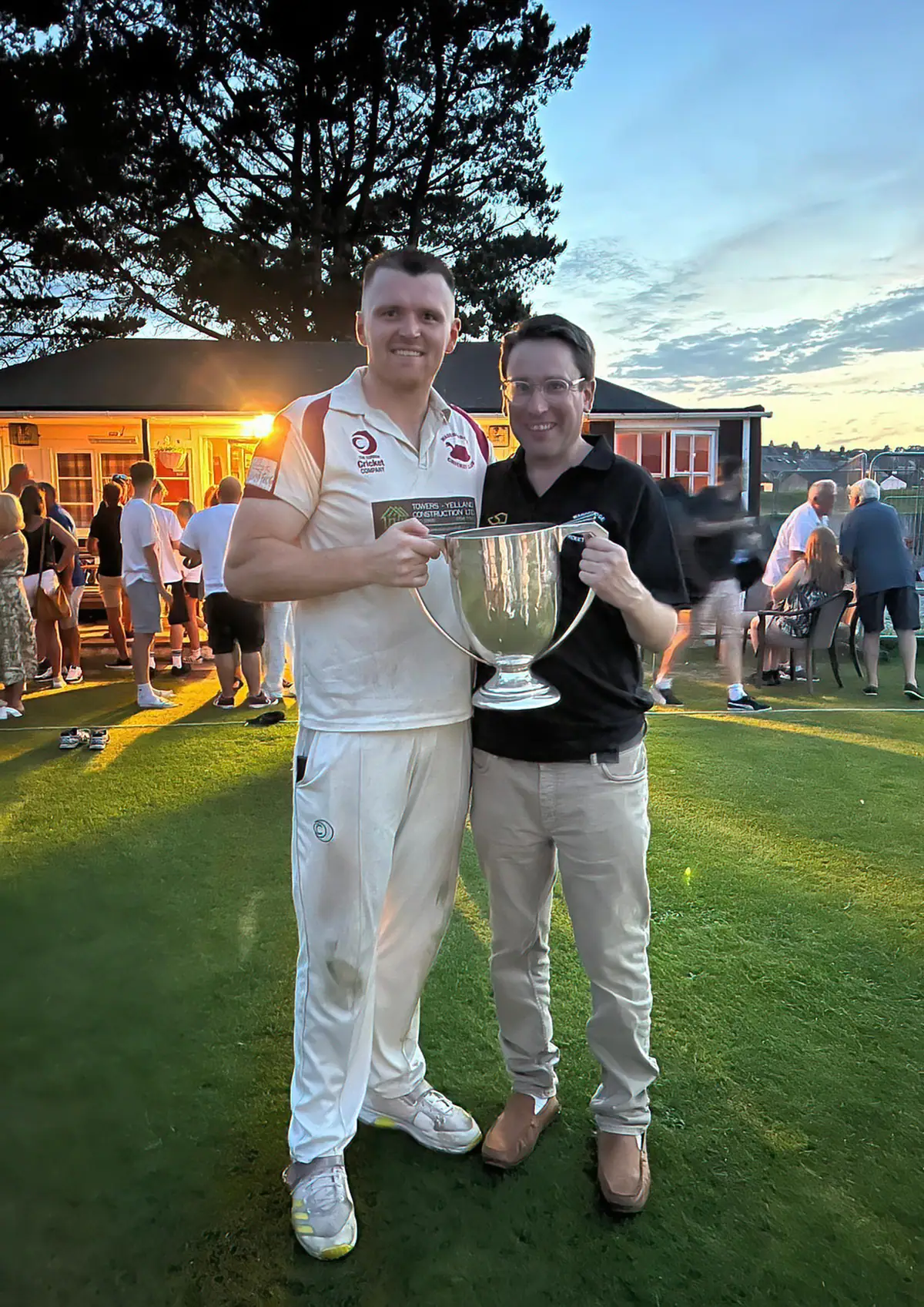 The Wadebridge captain in whites holds the championship cup with a club official as the sun goes down behind the clubhouse.