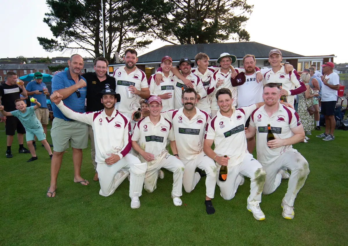 The 2023 team celebrate with supporters and families on the outfield, bottles raised, a clubhouse and trees behind.