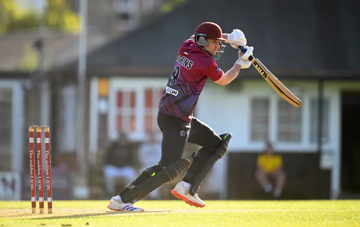 A Wadebridge batter in the claret and black kit drives off the front foot in evening sun, the white pavilion with its red window frames behind him.