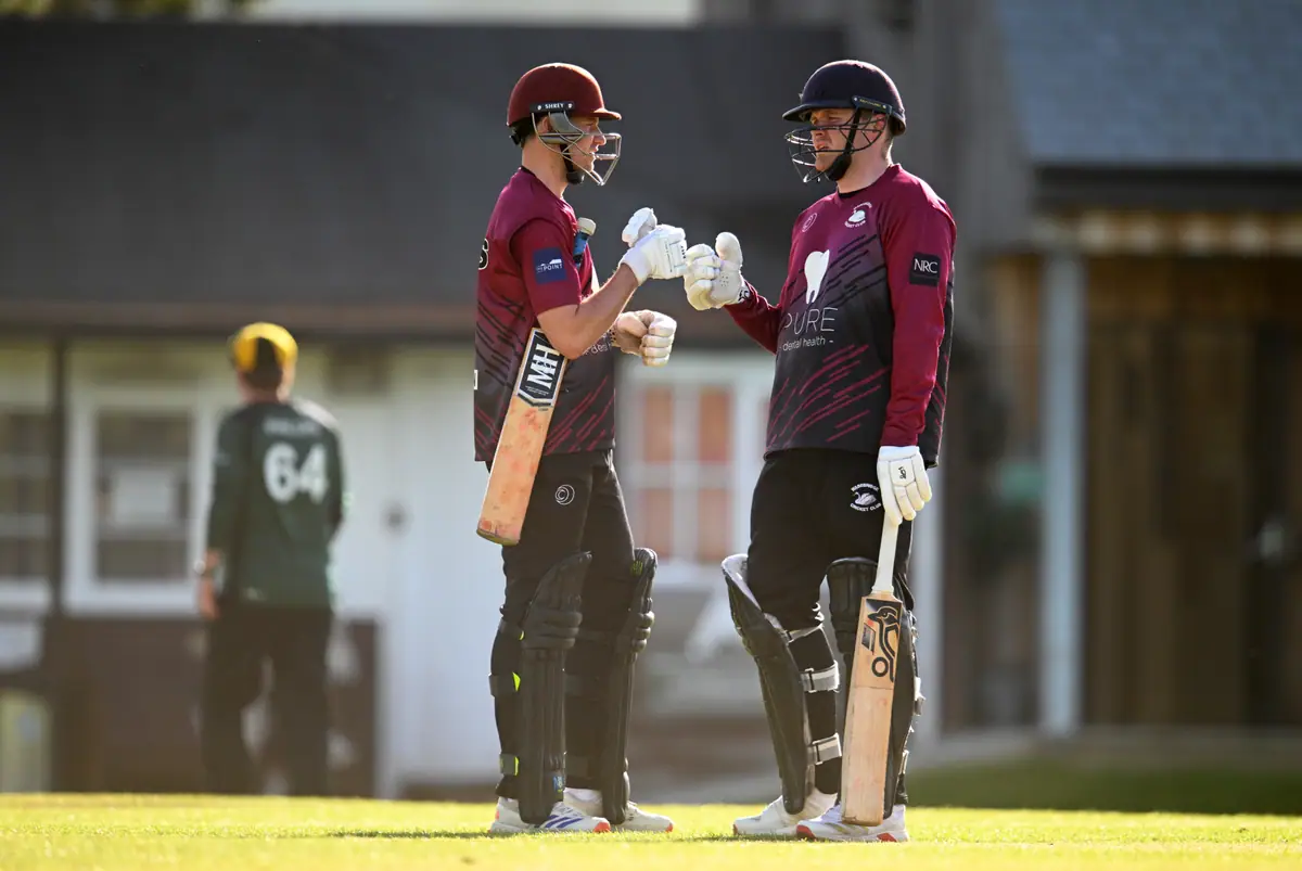 Two Wadebridge batters touch gloves mid-pitch in front of the timber pavilion.