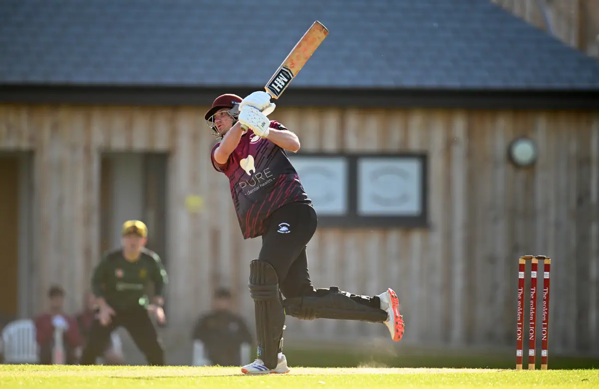 A Wadebridge batter sets off for a run in front of the timber pavilion, fielders watching.