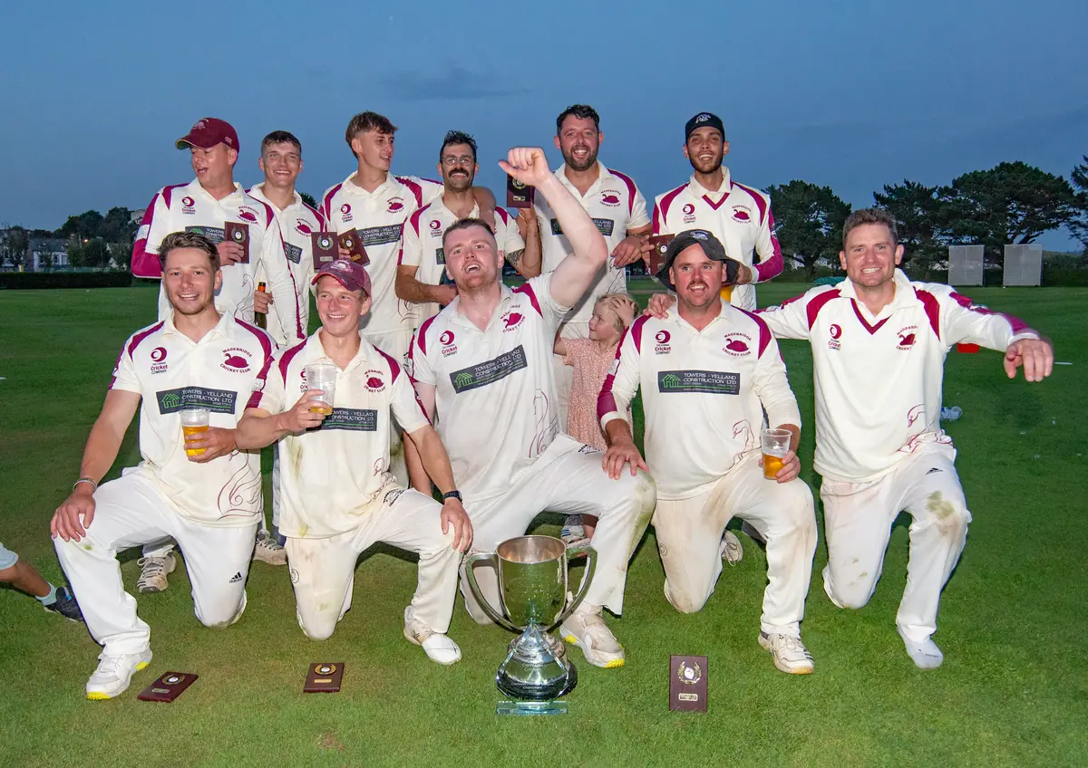 The 2023 Wadebridge 1st XI kneel at dusk around the County Championship trophy, one player punching the air.